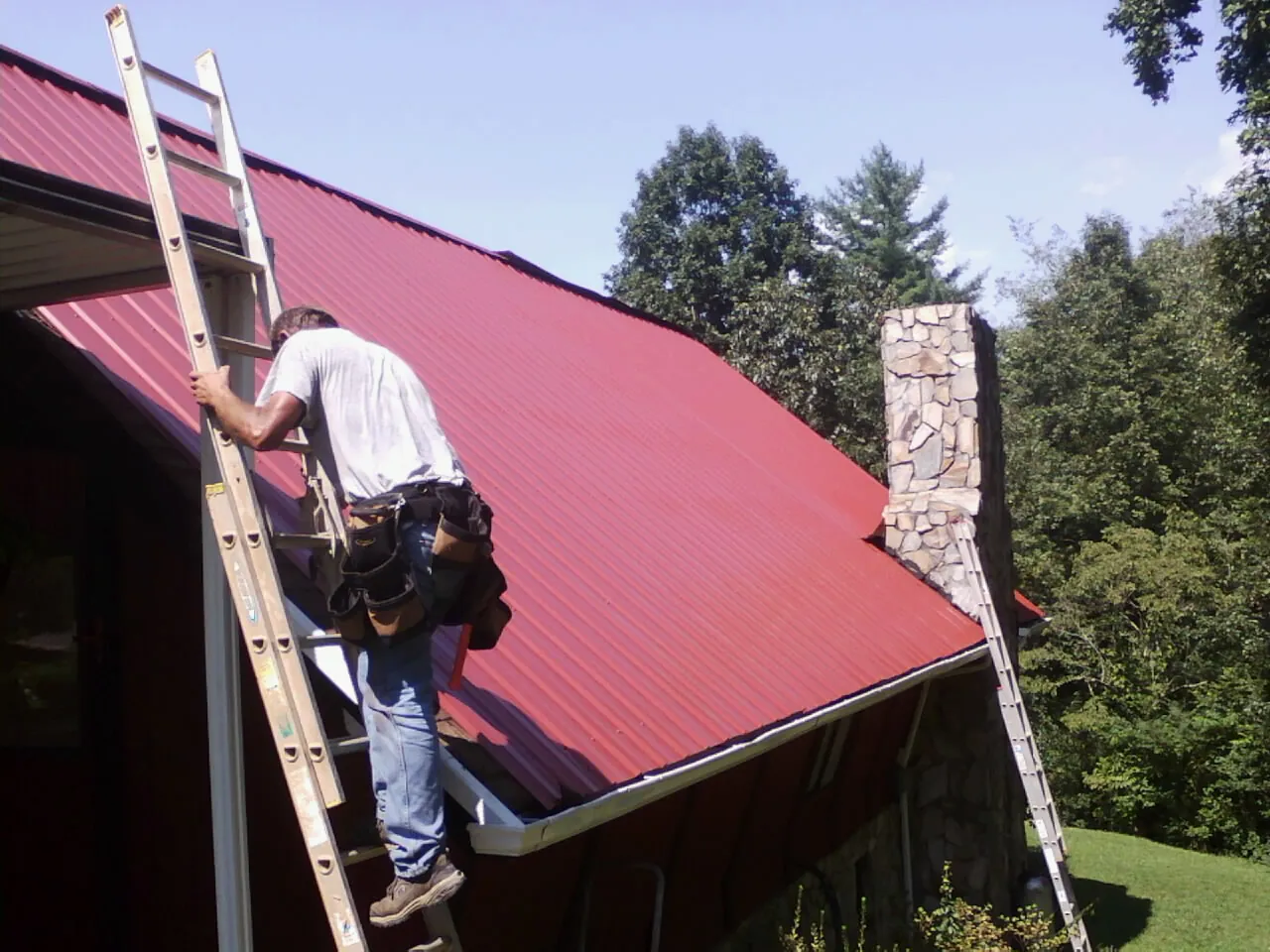 Roofer on a ladder beside a red metal roof and stone chimney — Soffit & Fascia Repair by Roofing Brothers in Copenhagen